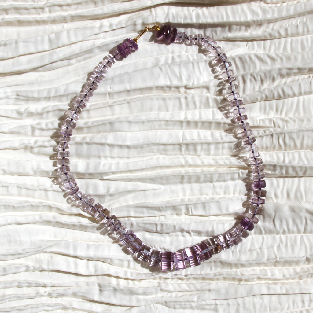 A beaded necklace made of faceted purple and lavender glass beads with a gold clasp, displayed on a textured white fabric background.