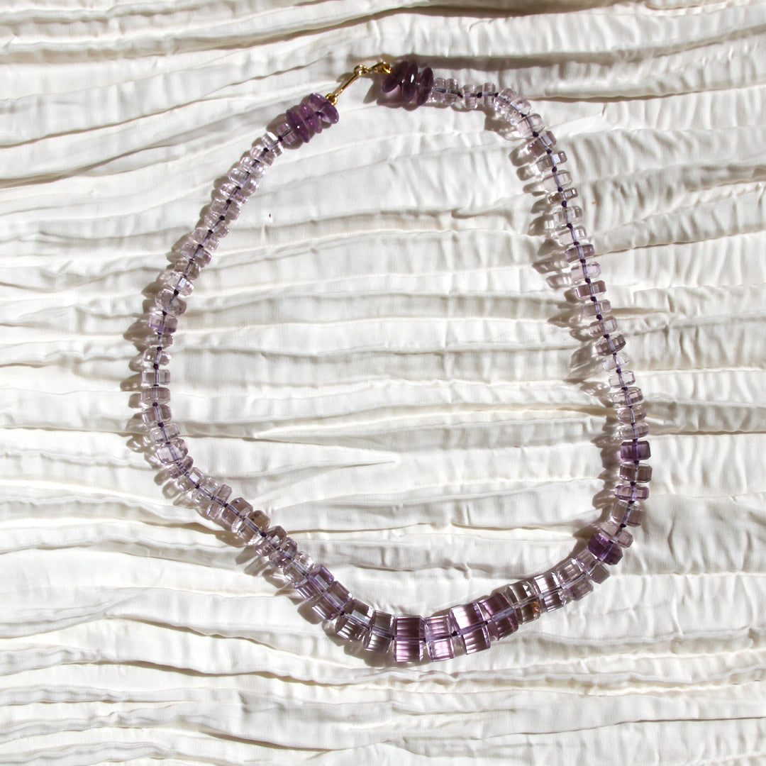 A beaded necklace made of faceted purple and lavender glass beads with a gold clasp, displayed on a textured white fabric background.