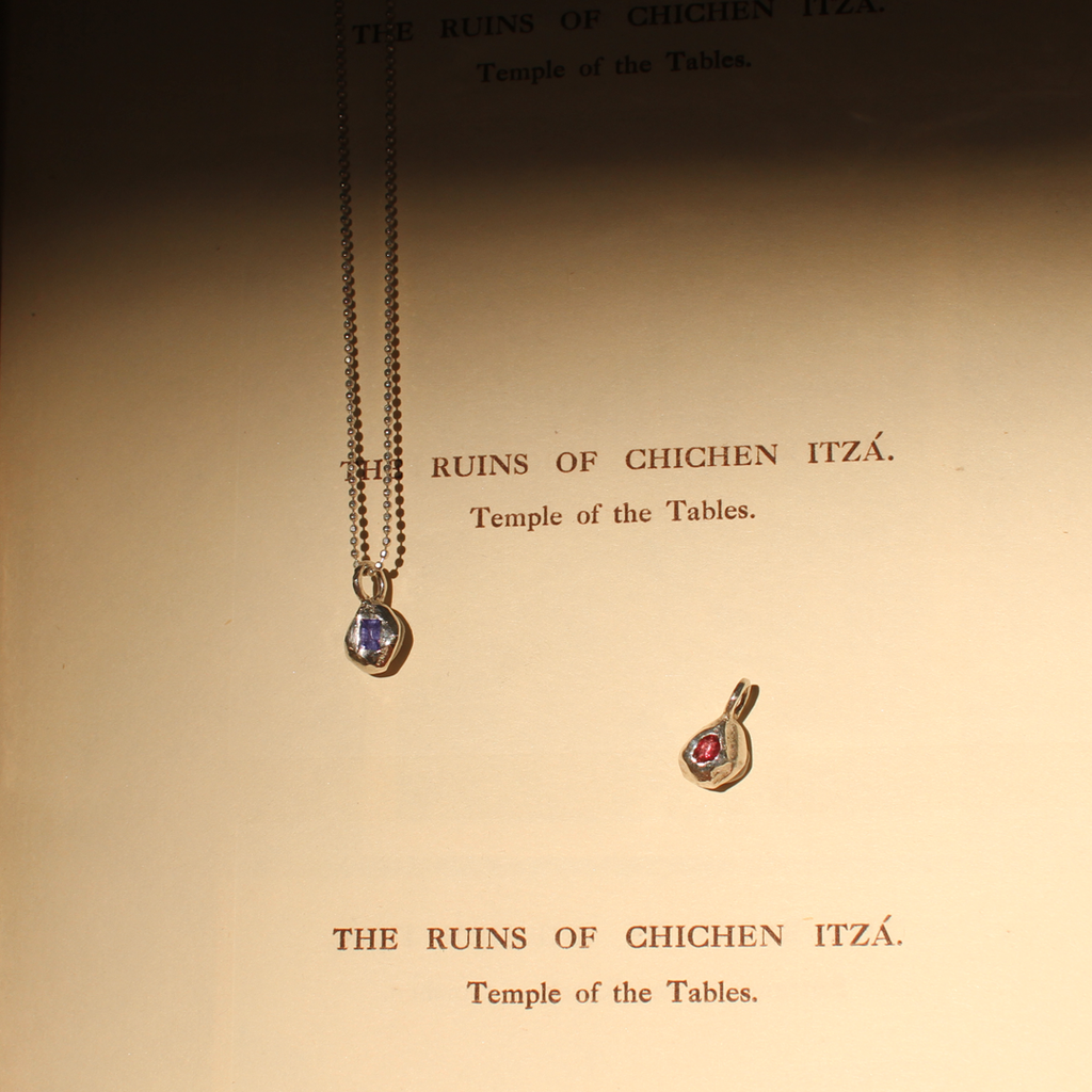 Two silver pendants with gemstones on a textured surface with text about Chichen Itza.
