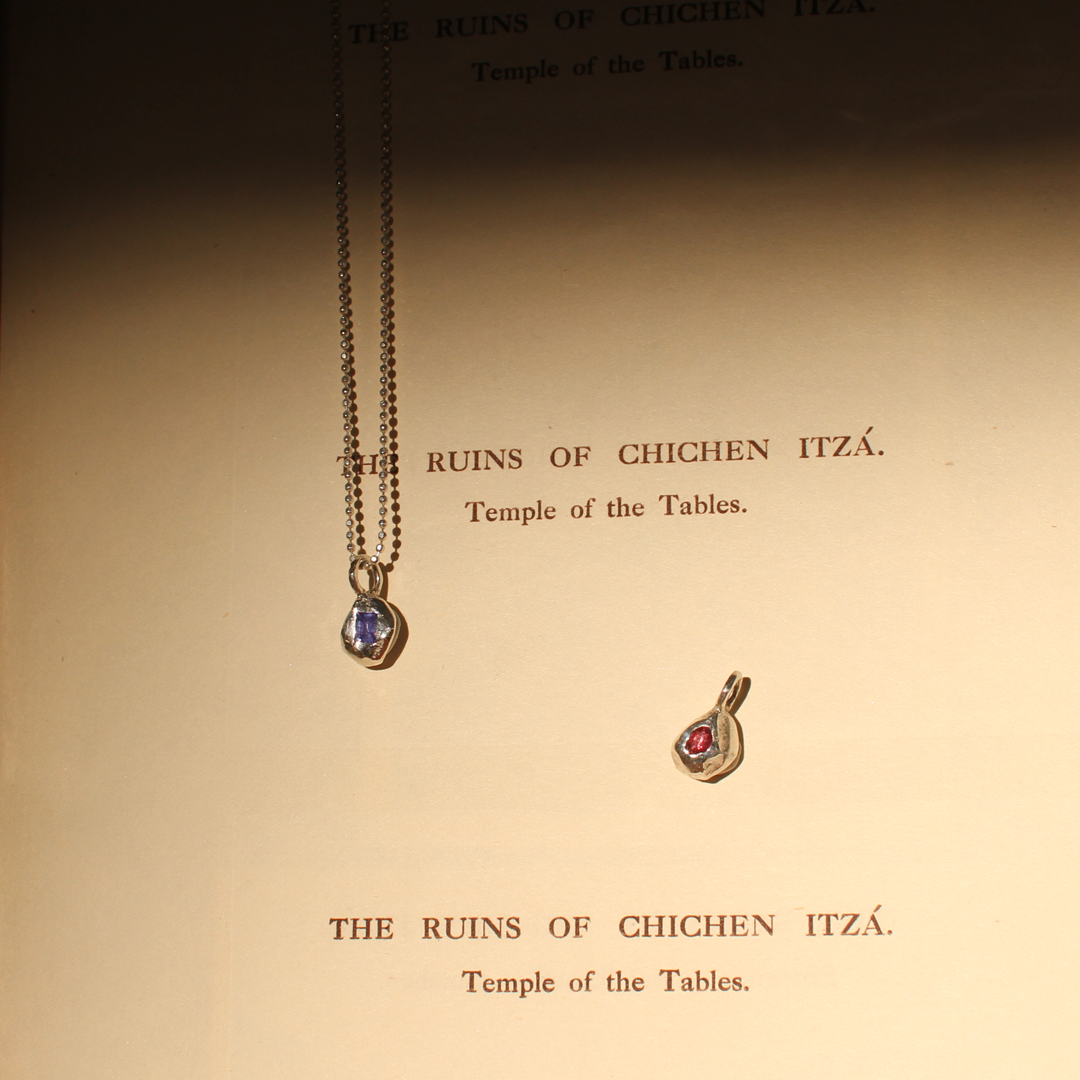 Two silver pendants with gemstones on a textured surface with text about Chichen Itza.